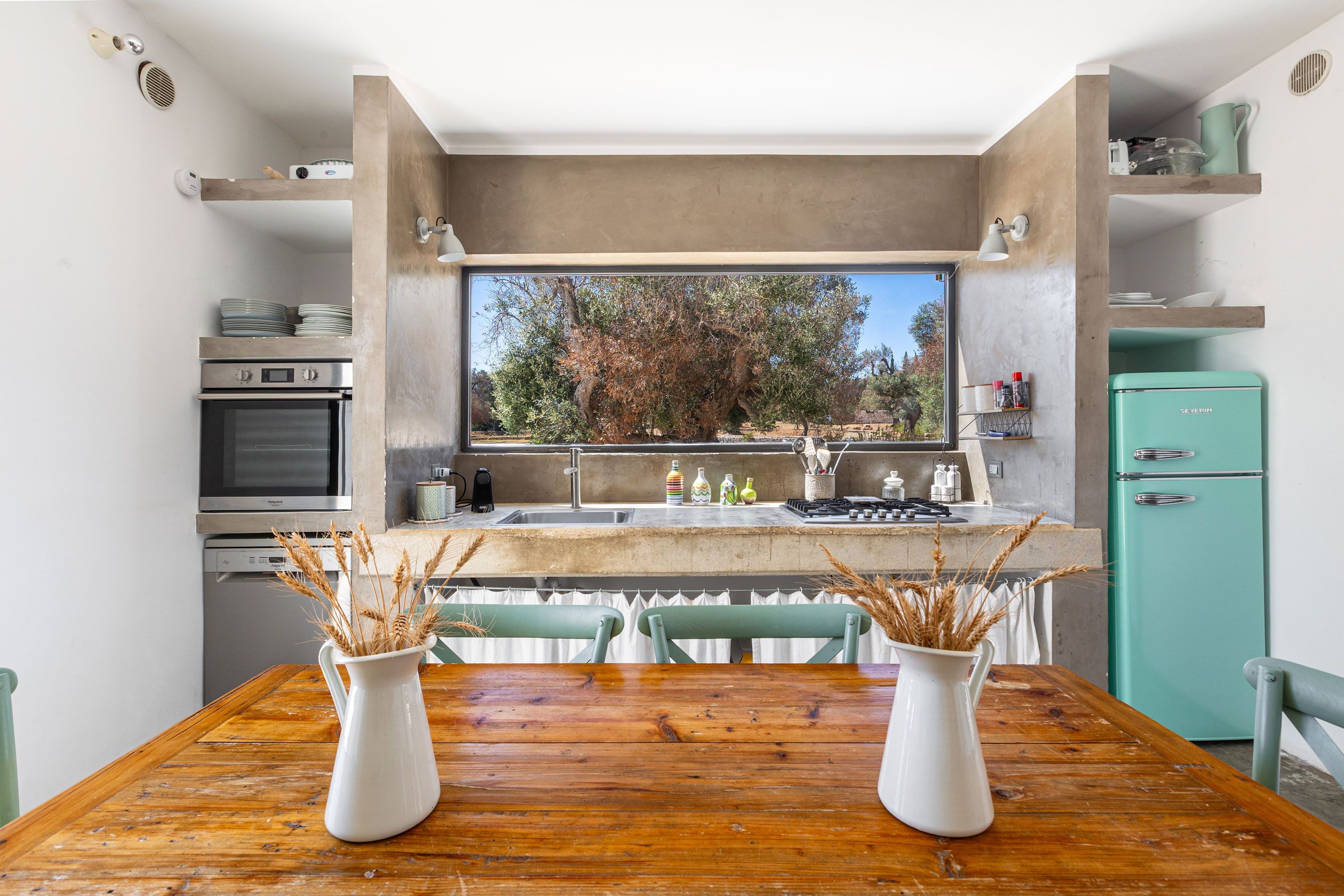 A kitchen with a dining table, two white vases with wheat stalks, a teal refrigerator, and a window overlooking trees.