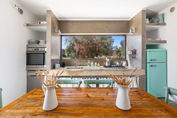 A kitchen with a dining table, two white vases with wheat stalks, a teal refrigerator, and a window overlooking trees.