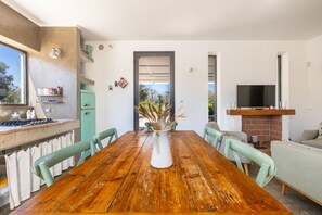 Ein Essbereich mit Holztisch, Stühlen, einer Vase mit Trockenpflanzen, eine Küche mit Herd und Kühlschrank sowie ein Fernseher auf einem Lowboard.