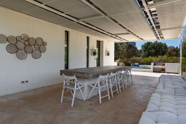 Un patio al aire libre con una larga mesa de comedor y sillas blancas, rodeado por un muro blanco con detalles decorativos circulares.