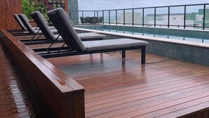 Terraza de madera con sillones de descanso y piscina, con vistas a un paisaje urbano.