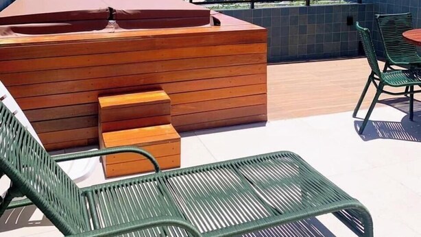 Un balcón con banca de madera y sillas verdes, con vista a una playa y al océano.