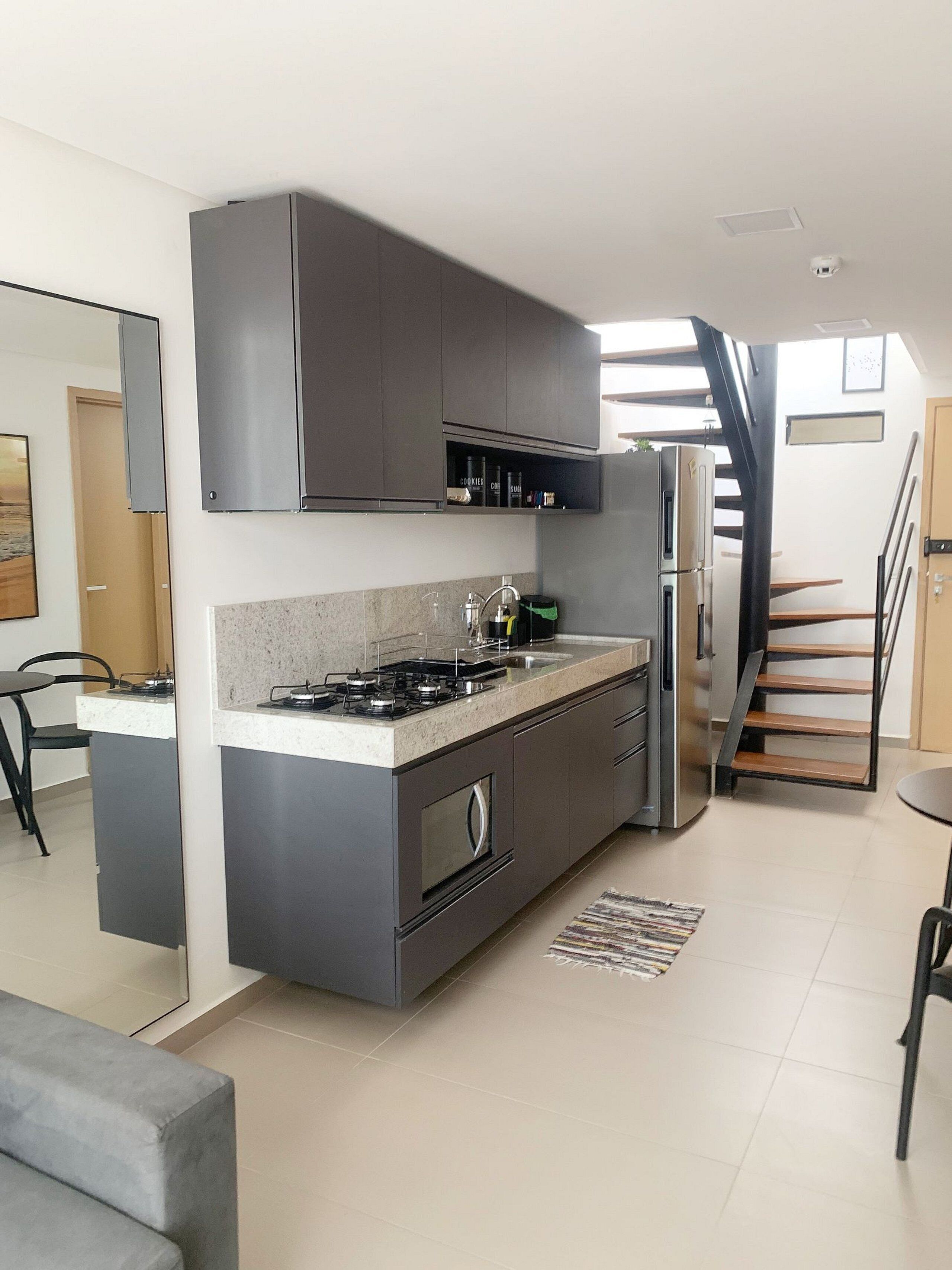 Una cocina moderna con armarios grises, una estufa empotrada y un refrigerador. Se ve una escalera que sube a otro nivel.