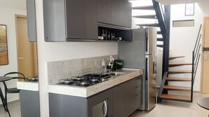 Una cocina moderna con armarios grises, una estufa empotrada y un refrigerador. Se ve una escalera que sube a otro nivel.