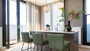A modern kitchen with a central island, three green bar stools, pendant lights, and a decorative plant. - Stylish Penthouse With 2 Bedrooms (The Hague)