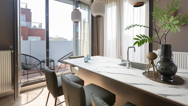 A modern kitchen with a large island, a dining area with chairs, and a view of the city through large windows. - Stylish Penthouse With 2 Bedrooms (The Hague)