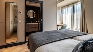 Una habitación de hotel moderna con una cama grande, un espejo, un espejo redondo, un baño con inodoro y lavamanos, y una ventana con cortinas.