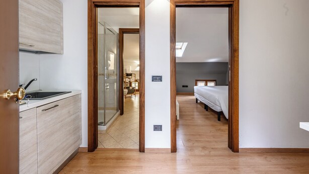 A modern kitchen with a sink and cabinets, a hallway leading to a bedroom with a bed and a nightstand, and a bathroom with a glass shower. - Emilia Studios 7 (Modena)