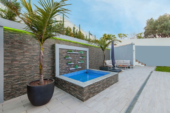 Un espace piscine extérieur moderne, agrémenté d’un mur en pierre, d’un jardin intégré avec un palmier, et d’un coin salon avec un canapé et des fauteuils.