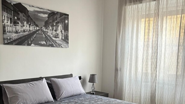A bedroom with a large bed, a nightstand, a lamp, a window with sheer curtains, and a cityscape painting on the wall.