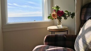 Une pièce avec une fenêtre donnant sur la mer, une petite table ronde ornée d’un vase de fleurs et un fauteuil à motifs écossais.