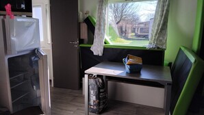 Una kitchenette compacta con refrigerador, microondas y un mueble pequeño. Hay una ventana con cortinas, una mesa con una silla y una mochila en el suelo.