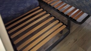 Una habitación pequeña y funcional con una cama individual, una mesita y un estante con objetos.
