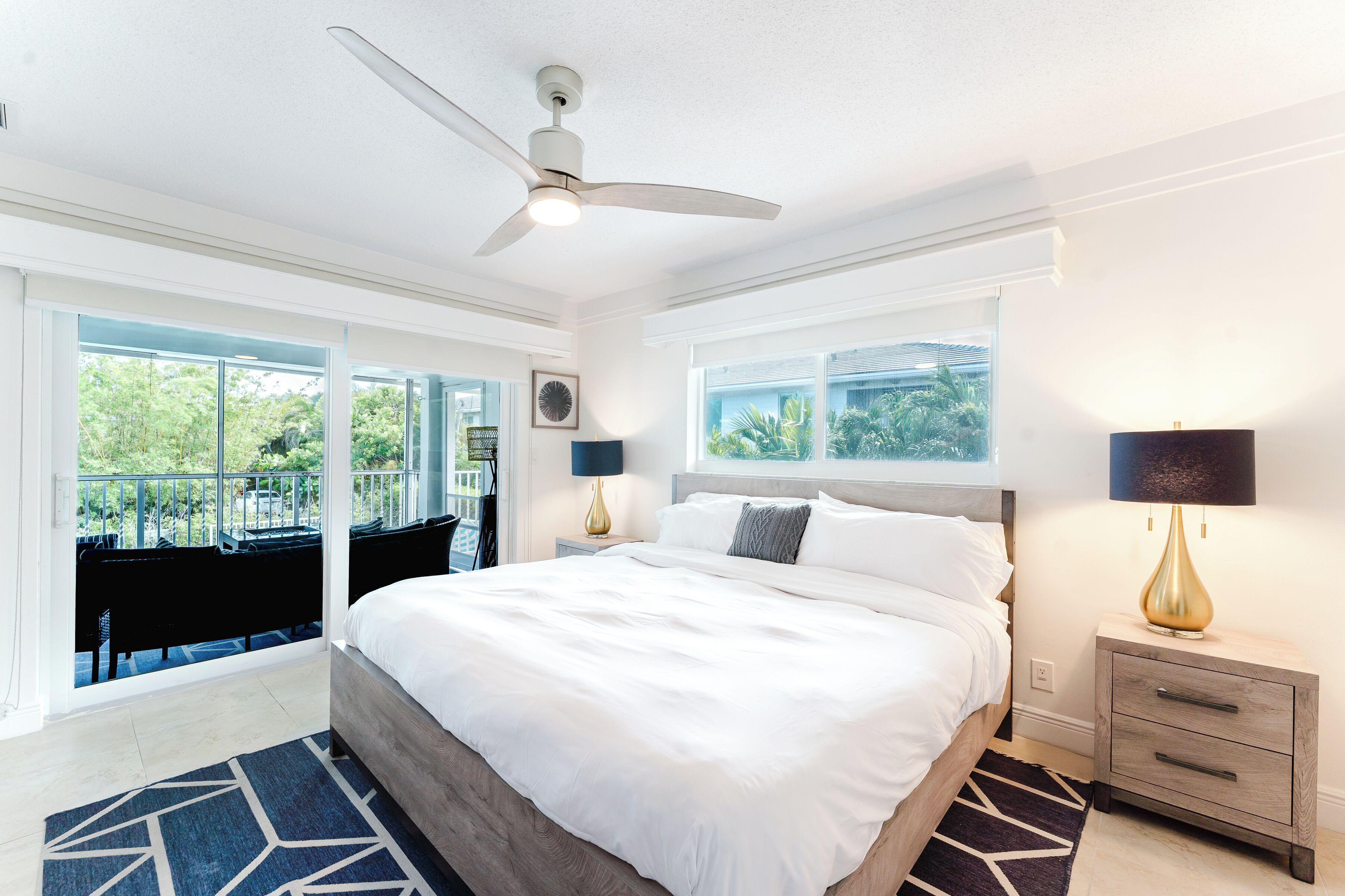A modern bedroom with a large bed, two bedside tables with lamps, a ceiling fan, and large windows offering a view of greenery.