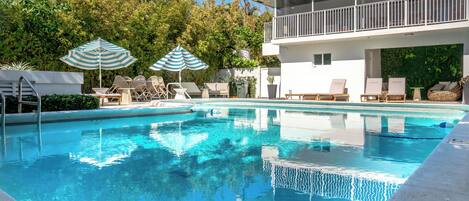 A swimming pool with lounge chairs and umbrellas, surrounded by greenery and a modern building with a balcony.