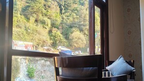 A room with a large window offering a view of a forested hillside and a stone wall. A wooden chair with a cushion and a pillow is positioned near the window. - The waypointinn (Shimla)