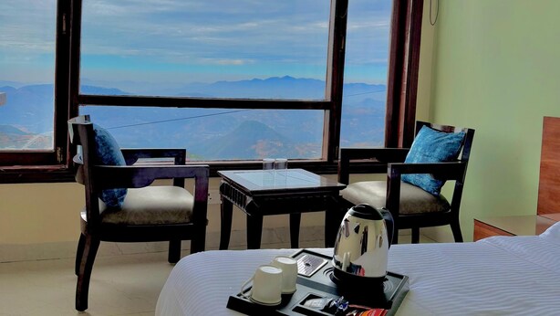 A hotel room with a large window offering a mountain view, a bed with a tray of breakfast items, and two chairs with blue cushions. - The waypointinn (Shimla)