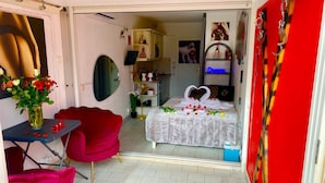 Une chambre avec un lit décoré de motifs en forme de cœur et de pétales de rose, une chaise rouge, une petite table sur laquelle est posé un vase de fleurs et un miroir au mur.