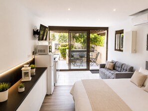 A modern bedroom with a bed, a sofa, a kitchen area, and a sliding glass door leading to an outdoor patio.
