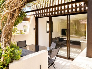 Une terrasse dotée d’une porte vitrée donnant sur un espace de vie, une table avec des chaises et un arbre recouvert de plantes grimpantes.