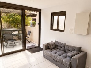 Un salon moderne avec un canapé gris, une porte vitrée donnant sur un espace extérieur équipé d’une table et de chaises, et une petite fenêtre.