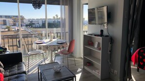 A modern living room with a dining area, large windows, a TV, and a balcony with a view of buildings. - Naturiste Cap d'Agde Héliopolis F 31 (Agde)