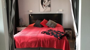 A modern bedroom with a large bed, grey curtains, a headboard, a bedside table, a lamp, and a wall art piece. - Naturiste Cap d'Agde Héliopolis F 31 (Agde)
