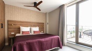 A modern hotel room with a large bed, a ceiling fan, a view of the city through large windows, and a balcony with outdoor furniture. - Spacious Apartment in Den Haag With View (The Hague)