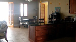 Una cocina amplia con una isla grande, armarios de madera e iluminación colgante. Hay un comedor con una mesa redonda y sillas, y un ventanal que proporciona luz natural.