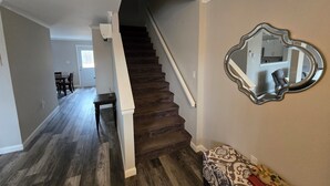 Un escalier avec des marches en bois foncé et une rampe blanche, menant à une pièce où se trouvent un pouf à motifs et un miroir au mur.