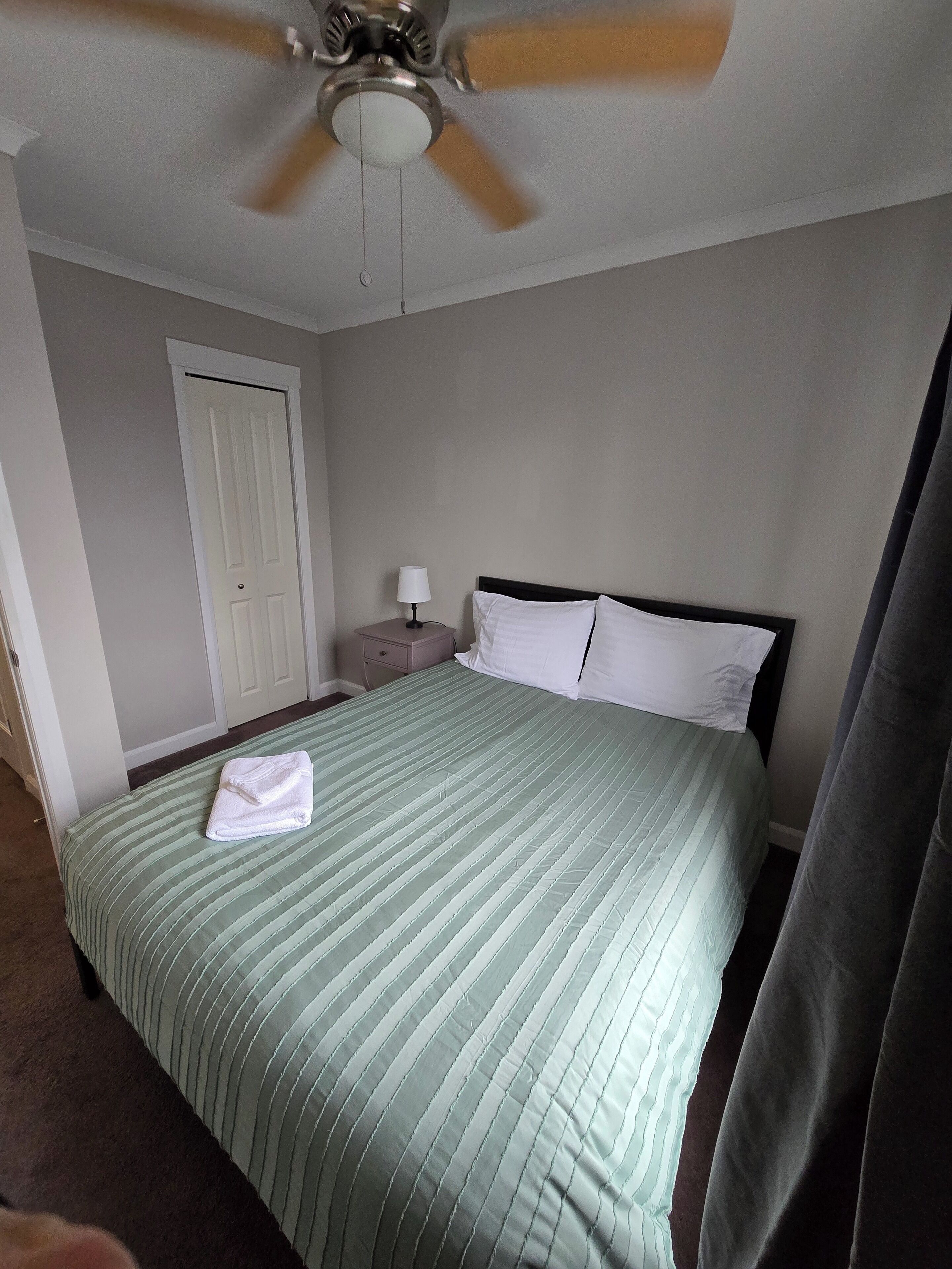 A hotel room with a bed, a nightstand, a lamp, a ceiling fan, and a door.