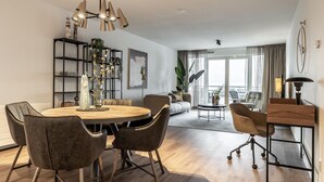 Una sala moderna con una mesa de comedor redonda de madera, sillas grises tapizadas, un sofá y una mesa de centro. Hay una estantería con objetos decorativos y un ventanal con cortinas.