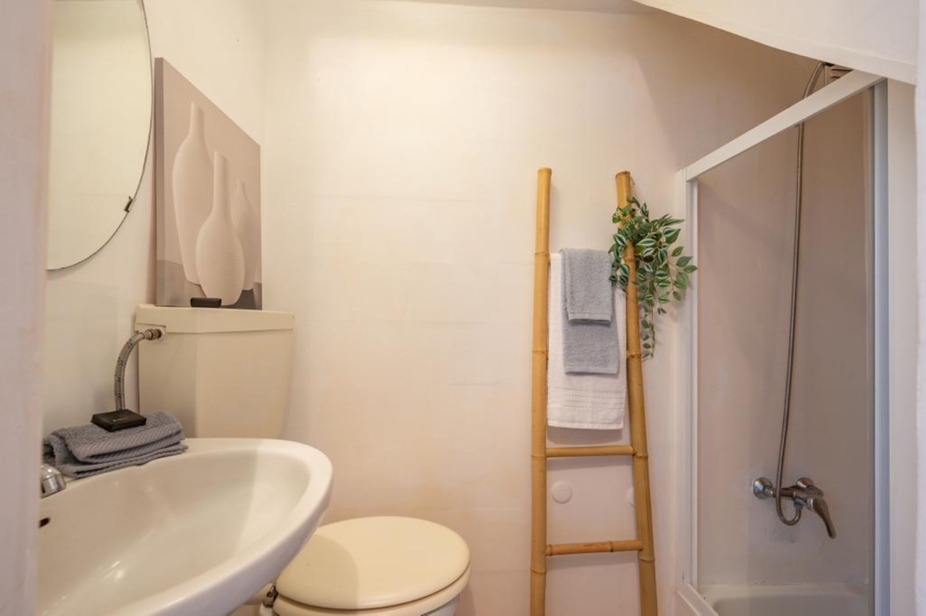 A bathroom with a white sink, toilet, and shower. There is a wooden ladder leaning against the wall and a plant on the shower curtain.