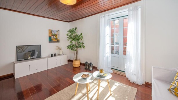 A living room with wooden flooring, a white sofa, a small table with a bowl, a potted plant, a TV on a white cabinet, and a window with white curtains. - Castle Fado River Alfama Flat (Lisbon)