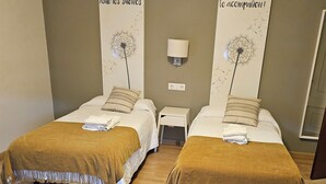 A hotel room with two beds, each with a brown blanket and pillows, a bedside table with a lamp, and a wall-mounted light. - HOSTAL MARIQUITO (Fisterra)