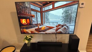 Televisor de pantalla plana montado en un mueble negro con detalles de chimenea, un florero con flores y una silla de mimbre.