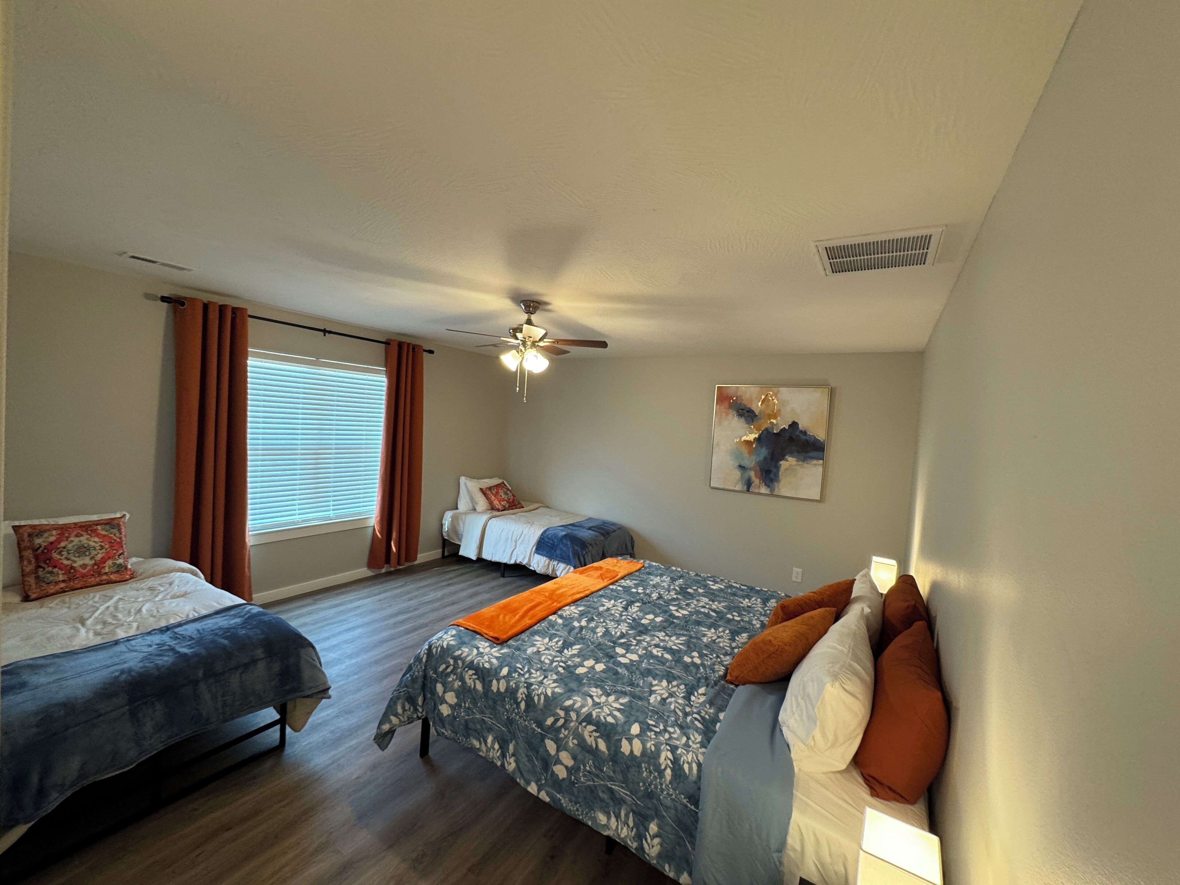 A bedroom with two beds, a ceiling fan, a window with curtains, and a painting on the wall.