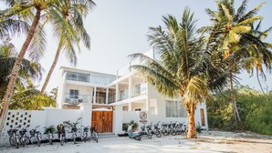 Un bâtiment moderne à la façade blanche, avec une porte en bois et une enseigne sur laquelle on peut lire « BELINA ». Des vélos sont garés devant et un palmier se trouve à droite.
