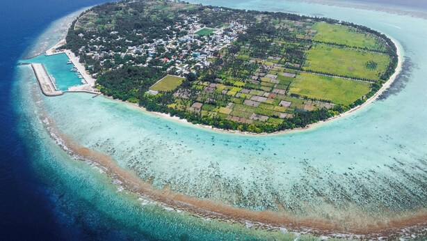 Une vue aérienne d’une île circulaire, entourée d’une mer d’un bleu profond, sur laquelle on aperçoit un quai, une plage de sable et un mélange de zones résidentielles et agricoles.
