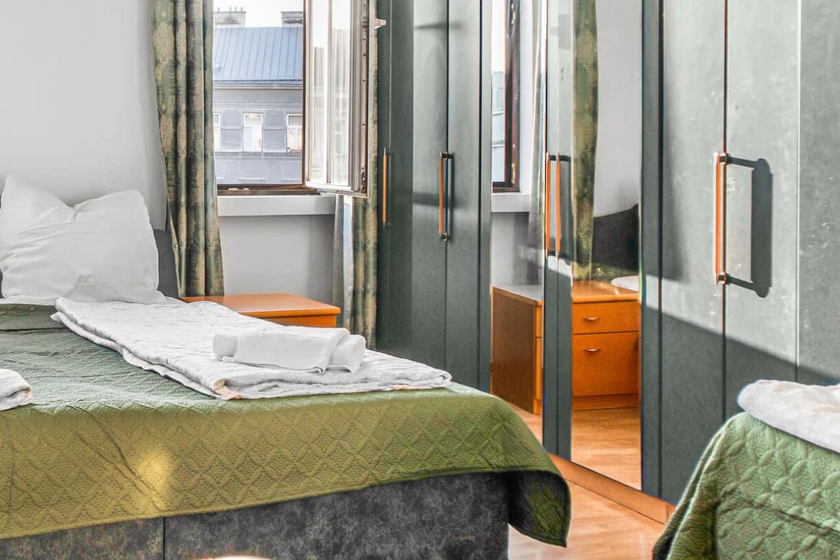 A neatly made bed with pillows and towels, a bedside table, a wardrobe, and a view of buildings through the window.