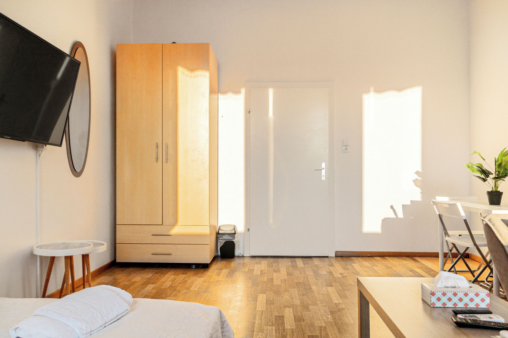 A hotel room with a bed, a TV mounted on the wall, a wooden wardrobe, a small table with a plant, and a desk with a chair.