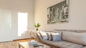 Una sala de estar con sofá, mesa de centro, un cuadro de un paisaje urbano, planta en maceta y una puerta.
