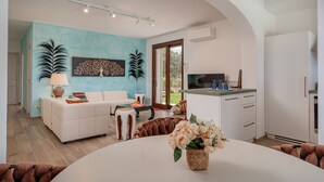 Una sala moderna con un sofá, una mesa de centro y almohadones decorativos. Hay un comedor con una mesa y sillas, y una cocina con encimera y armarios.