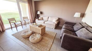 Een woonkamer met een bank, een bijzettafeltje en een stoel. Er is een raam met gordijnen en uitzicht op een gazon.