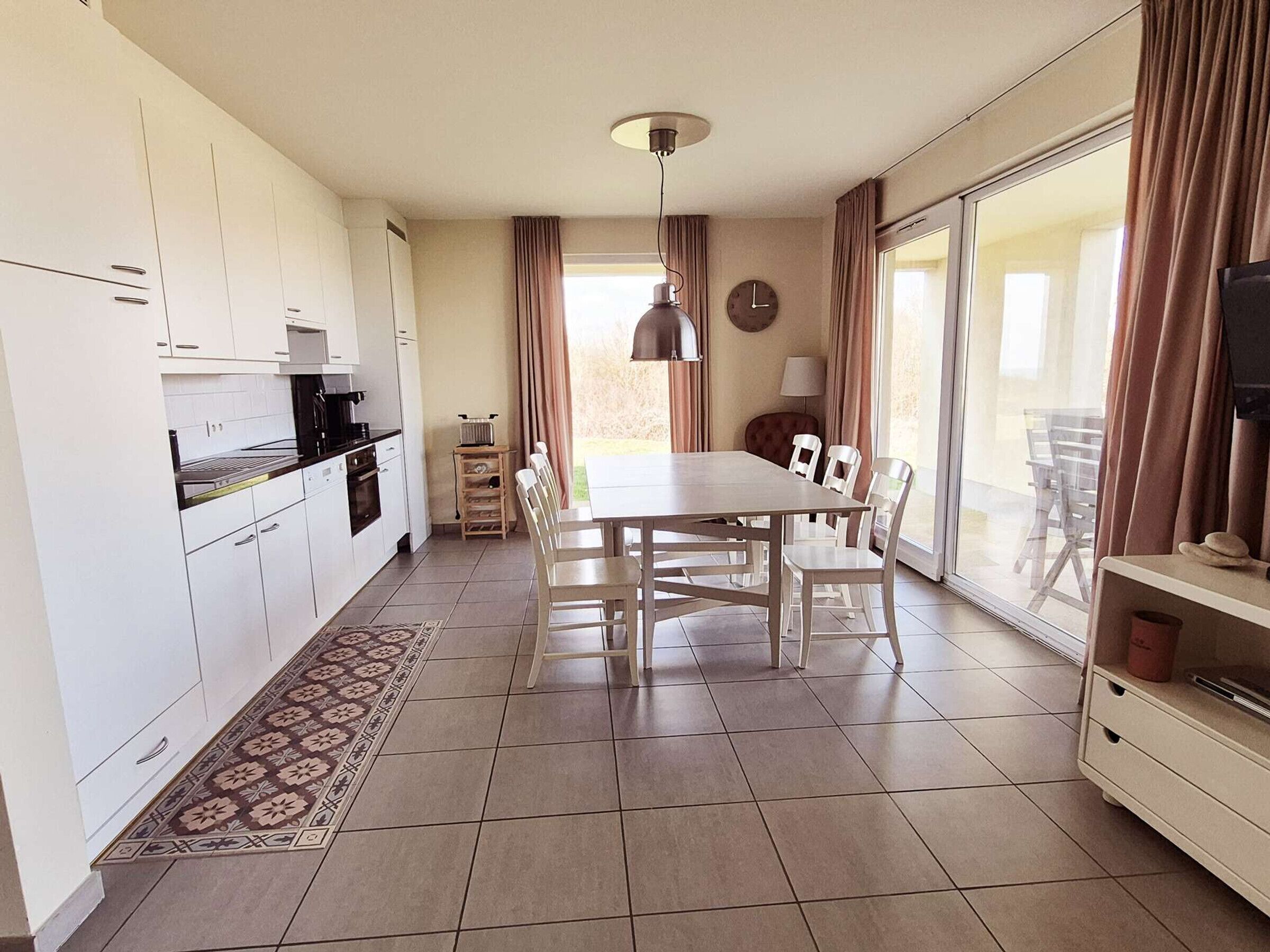 Een moderne keuken met witte kastjes, een eettafel met stoelen, een vloerkleed met patroon en een groot raam met gordijnen.