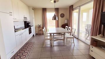 Een moderne keuken met witte kastjes, een eettafel met stoelen, een vloerkleed met patroon en een groot raam met gordijnen.