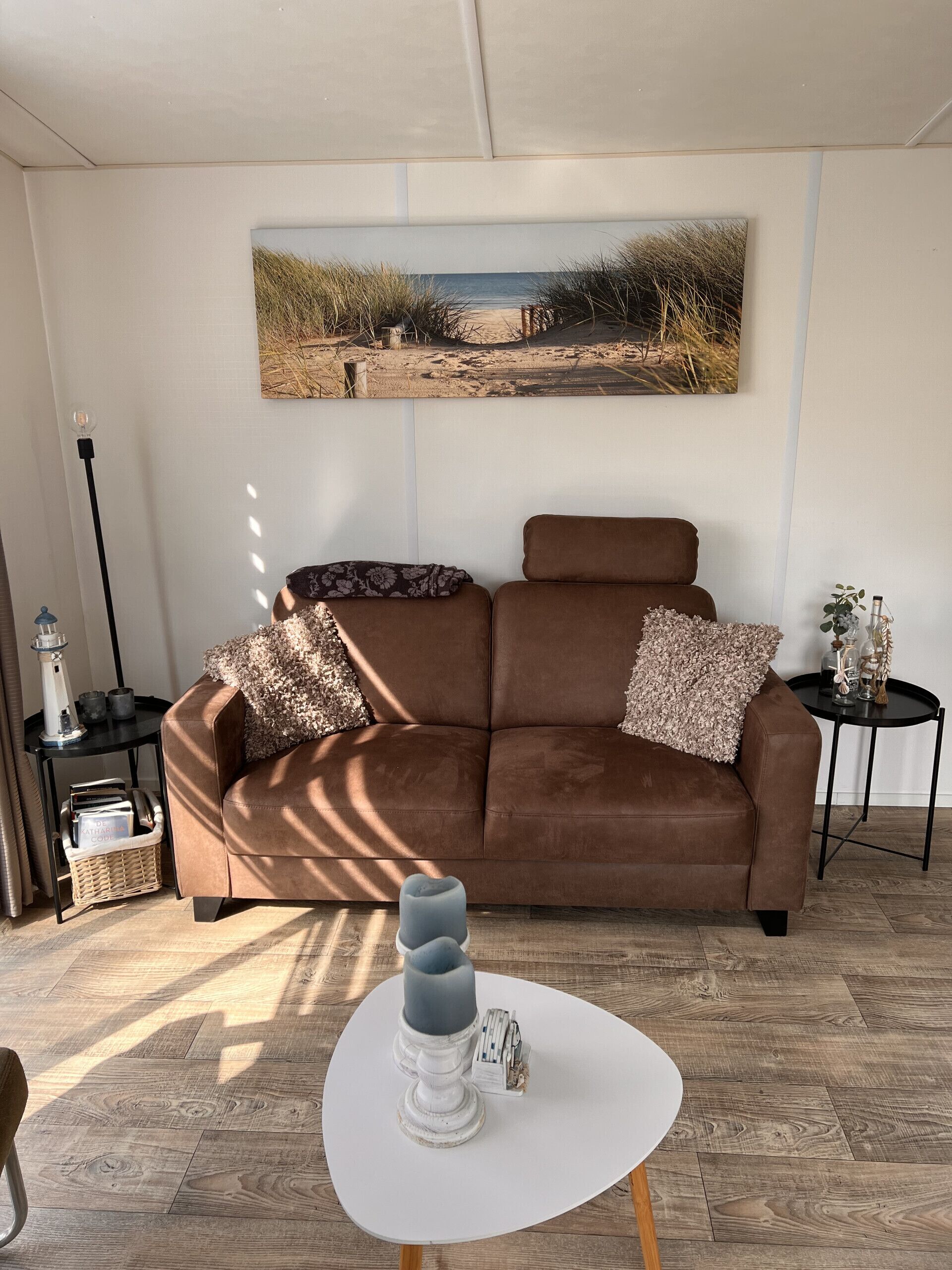 Een woonkamer met een bruine bank, een witte tafel met kandelaar, en een schilderij van een strandgezicht aan de muur.