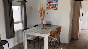 A dining area with a wooden table, chairs, a vase with dried plants, a painting on the wall, and a window with curtains.