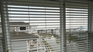 A view through window blinds of a residential area with houses, a body of water, and a bridge. - Relax in Style Oceanview Villa w/ Resort Amenities & Steps to the Beach (Ocean Isle Beach)