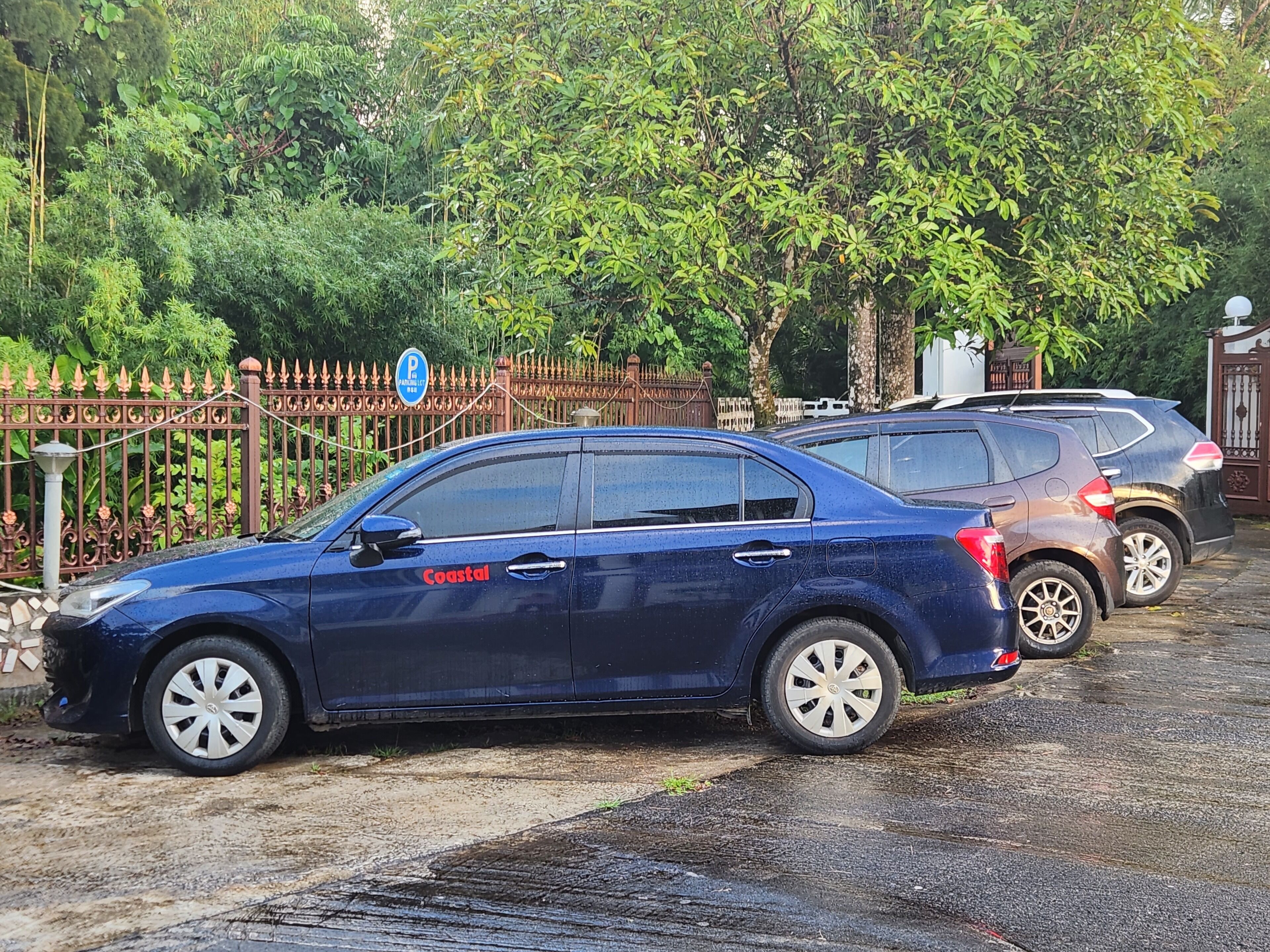 A blue car parked in a driveway with a metal fence and greenery in the background.